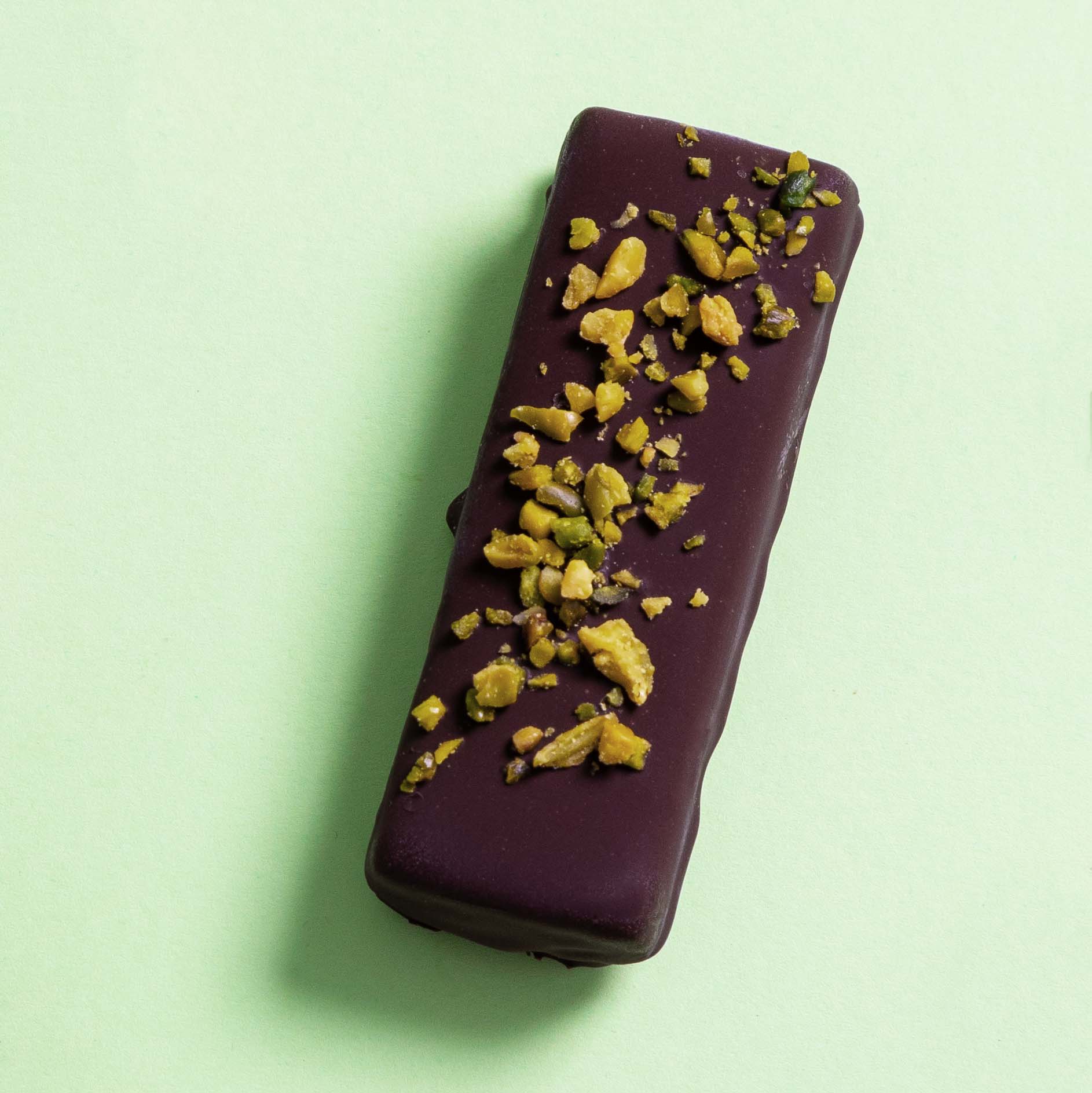 Ein zuckerfreier veganer Eis-Riegel mit einer dunklen Schokoladenhülle, der mit gehackten Pistazienstücken bestreut ist. Der Eisriegel liegt auf einer hellgrünen Oberfläche und ist eine ideale kalorienarme Snackoption