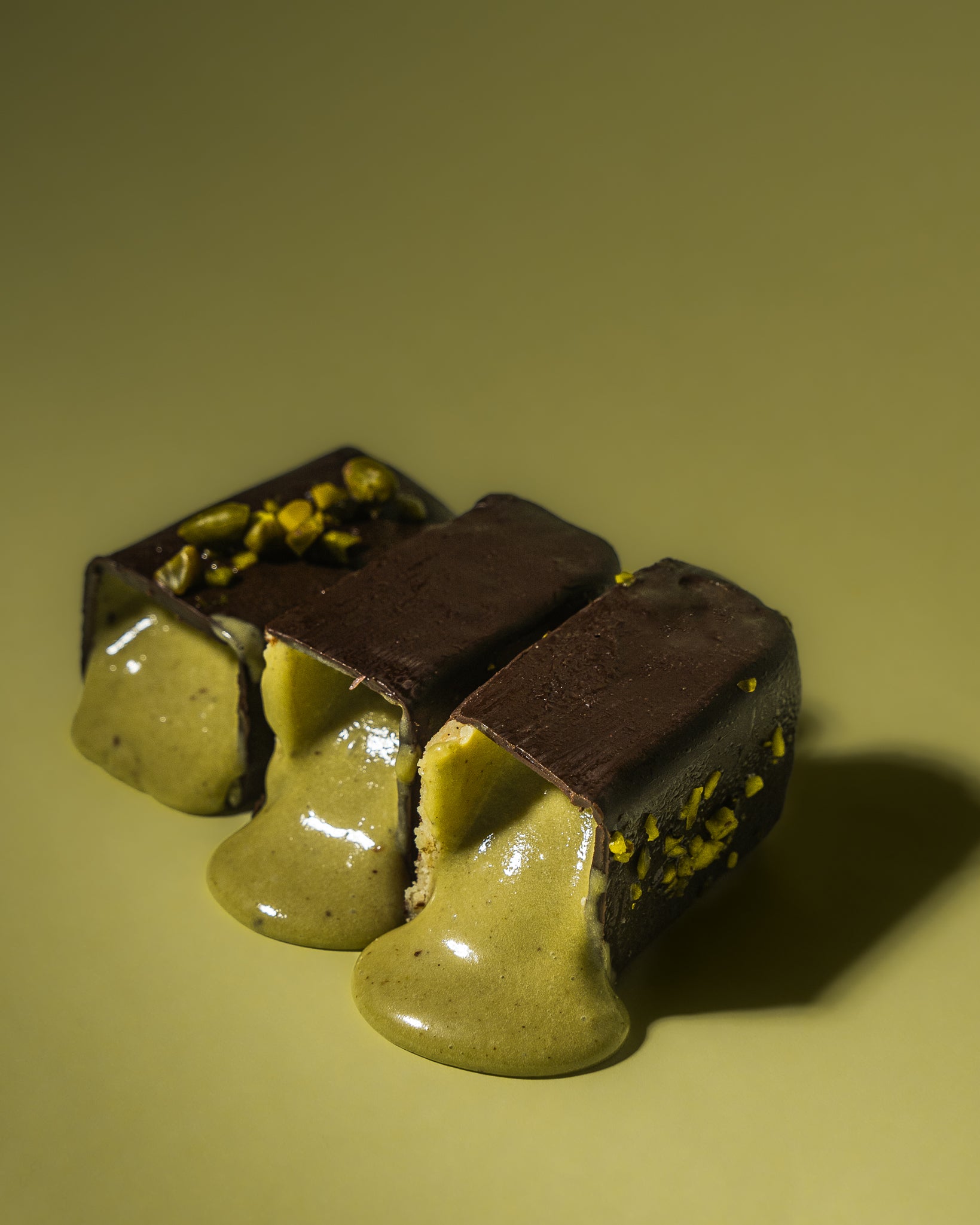 Drei aufgeschnittene Frozen Power  Eis-riegel mit dunkler Schokoladenhülle und flüssiger Pistazienfüllung, die appetitlich aus dem Inneren herausfließt. Oben sind kleine Pistazienstücke zu sehen. Der Snack ist zuckerfrei und vegan, perfekt für Fitness-Fans, die nach  und gesunden Alternativen suchen