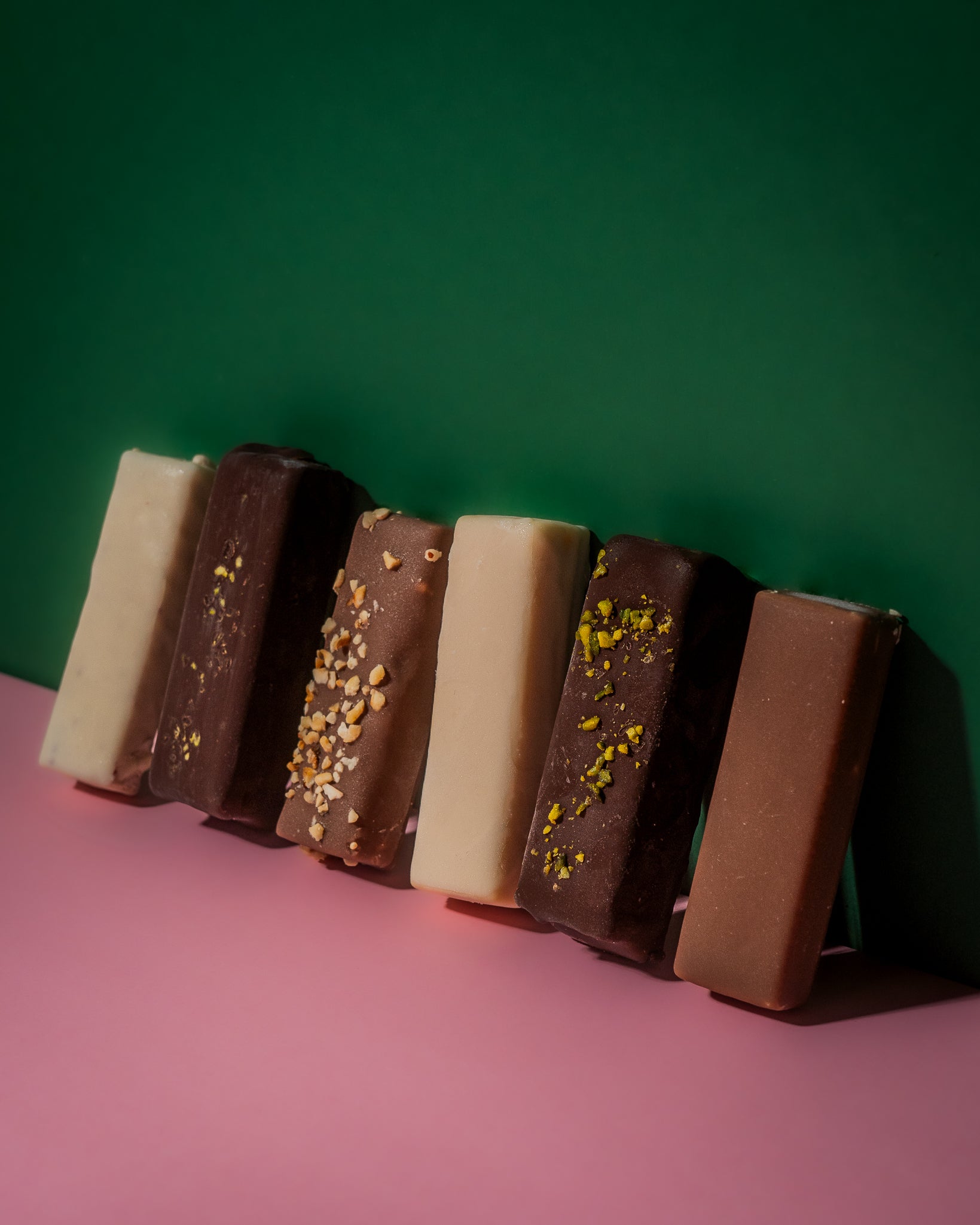 Eine Reihe von Protein und Veganen Eis-Riegeln von Frozen Power in verschiedenen Geschmacksrichtungen, die schräg auf einer rosa Oberfläche und vor einem grünen Hintergrund aufgereiht sind.