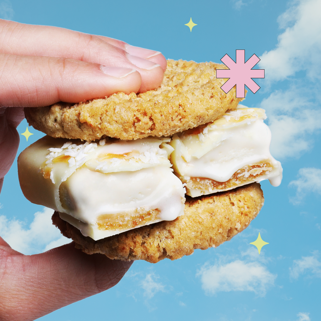 Eine Hand hält einen Eissandwich bestehend aus zwei Haferkeksen, und in der Mitte einen halbierten Protein-Eisriegel ohne Zuckerzusatz in der Sorte Creamy Coco. Der Hintergrund zeigt einen blauen Himmel mit ein paar Wolken.