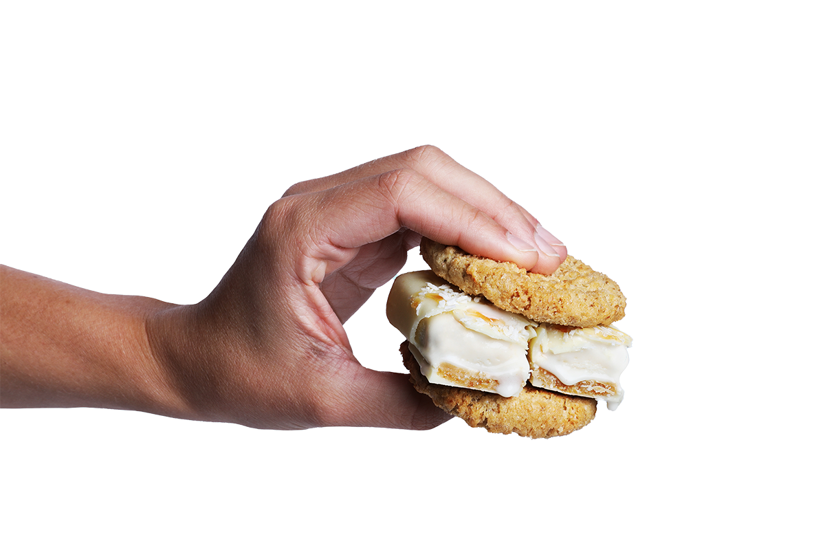 Eine Hand hält einen Cookie-Sandwich, bestehend aus zwei knusprigen Keksen und einer cremigen Füllung aus Protein-Eis. Die Füllung besteht aus einem High Protein Eis-Riegel von Frozen Power in der Sorte Creamy Coco