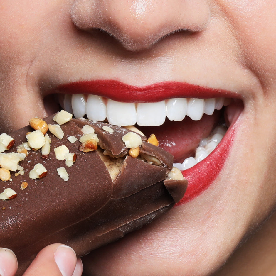 Frau beißt in einen mit Nüssen bestreuten Protein Eis-Riegel in der Sorte Crispy Hazel von Frozen Power mit Schokoladenüberzug, idealer kalorienarmer Fitness-Snack ohne Zuckerzusatz.