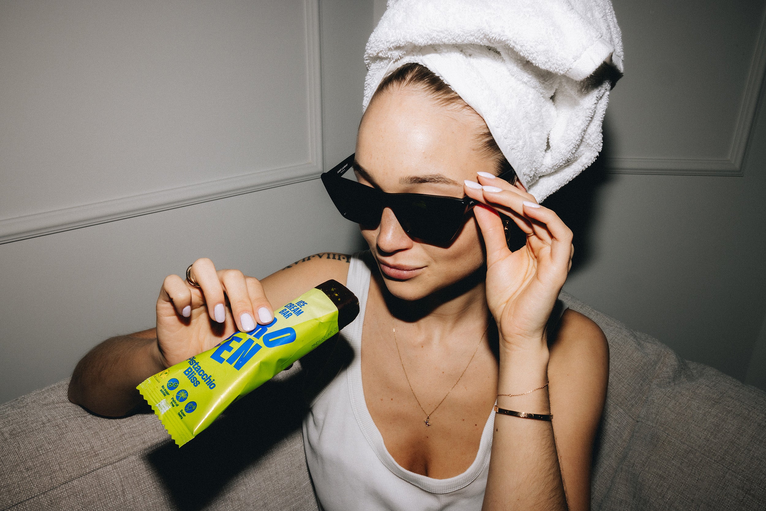 Frau mit Handtuch auf dem Kopf und Sonnenbrille hält einen Pistachio Bliss Eisriegel in der Hand. Sie trägt lässige Kleidung und genießt ihren Moment der Entspannung.