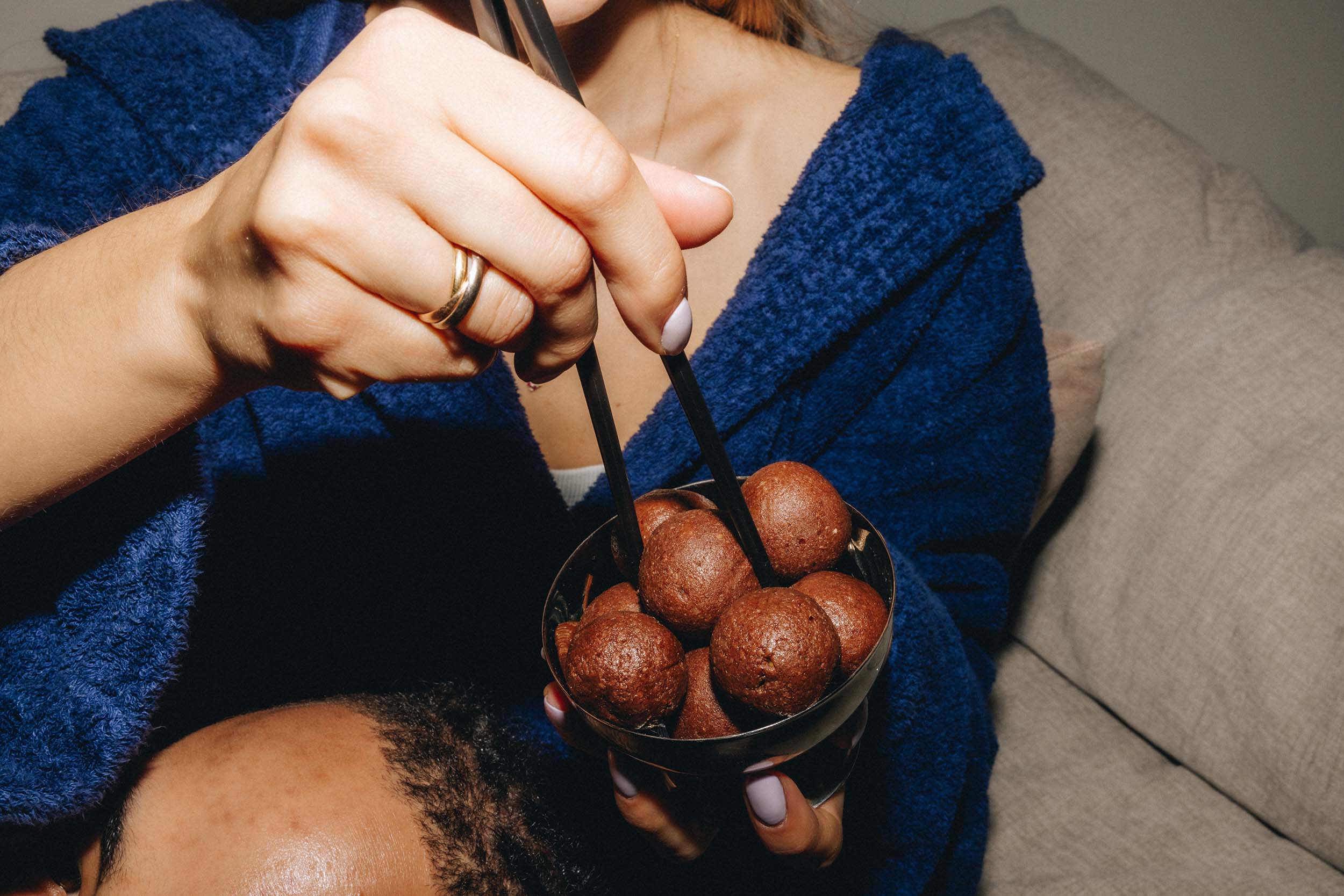 Nahaufnahme einer Hand, die mit Stäbchen die veganen Dattel Energy Bällchen von Frozen Power aus einer Schüssel hebt. Die Person trägt einen blauen Bademantel und genießt einen gesunden Snack.