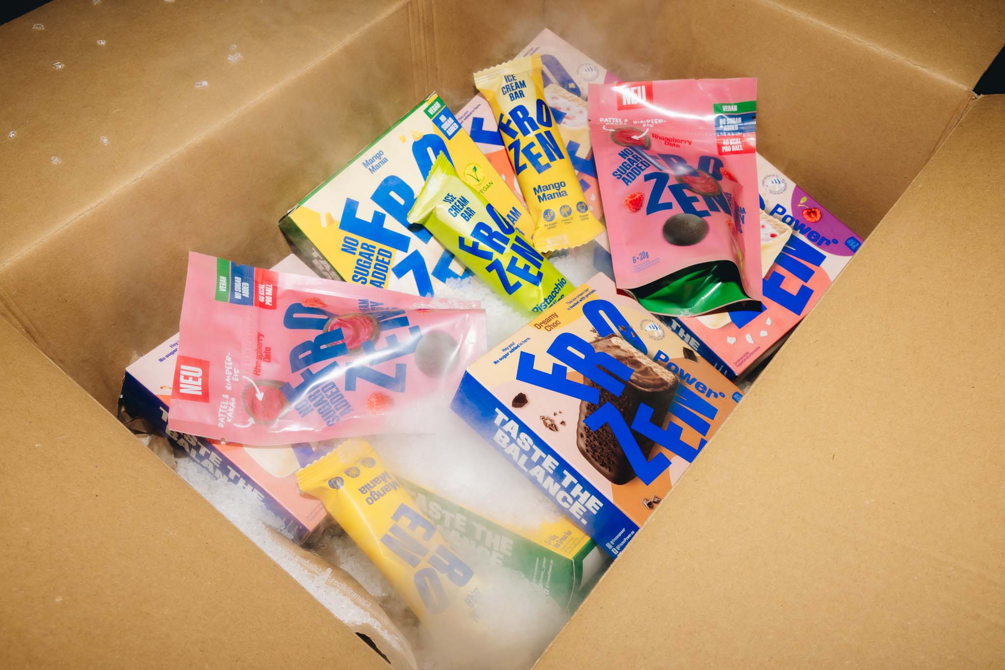 Ein Karton gefüllt mit verschiedenen Verpackungen von zuckerfreien und veganen Frozen Power Produkten. Die Produkte betonen eine gesunde, kalorienarme Ernährung und sind ideal als Fitness-Snacks und Protein-Snacks für Muskelaufbau geeignet