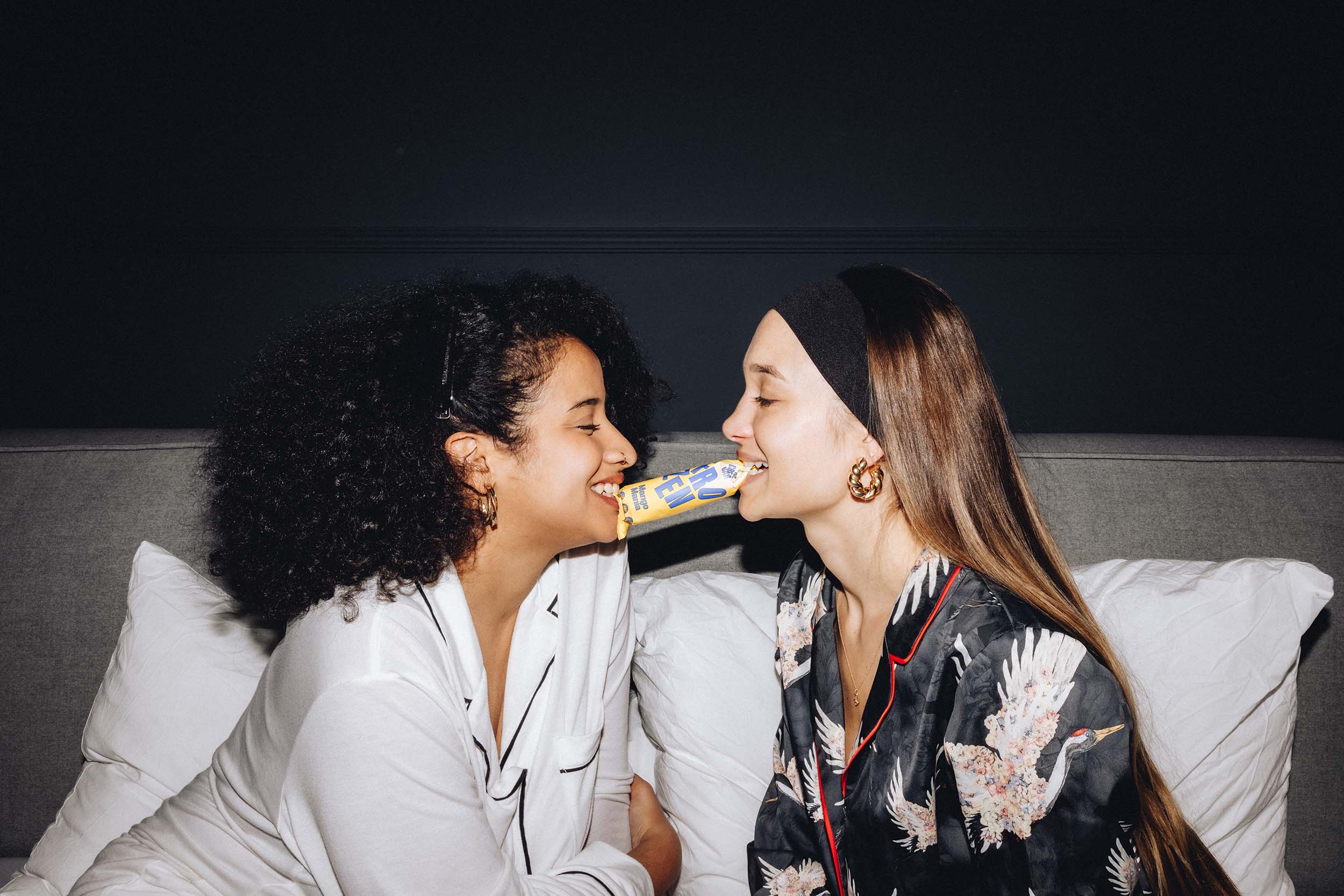 Zwei Frauen teilen sich einen veganen Mango Eis-Riegel. Der Moment betont gesunde Fitness-Snacks und die Freude am gemeinsamen Genuss zuckerfreier, proteinreicher Leckereien