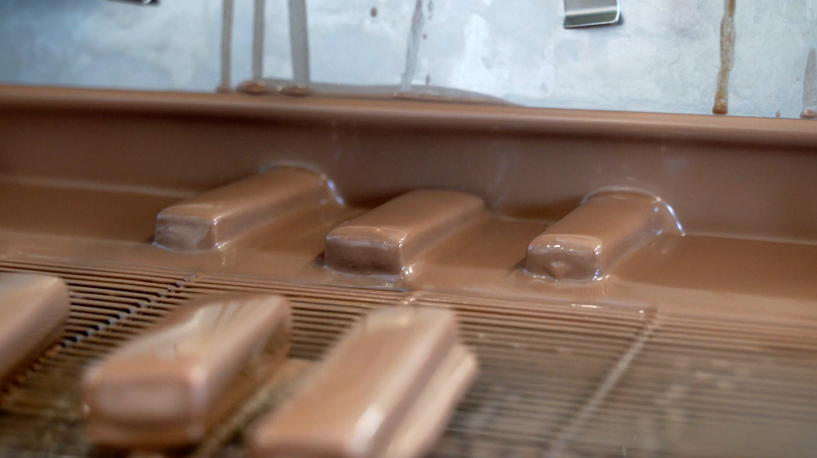 Ein Produktionsprozess zeigt Protein-Eisriegel von Frozen Power, die in eine Schokoladenschicht getaucht werden. Die Eisriegel bewegen sich über ein Förderband und werden vollständig mit einer cremigen Schokoladenhülle überzogen. Dieser Schritt ist Teil der Herstellung von proteinreichen, zuckerfreien Snacks.