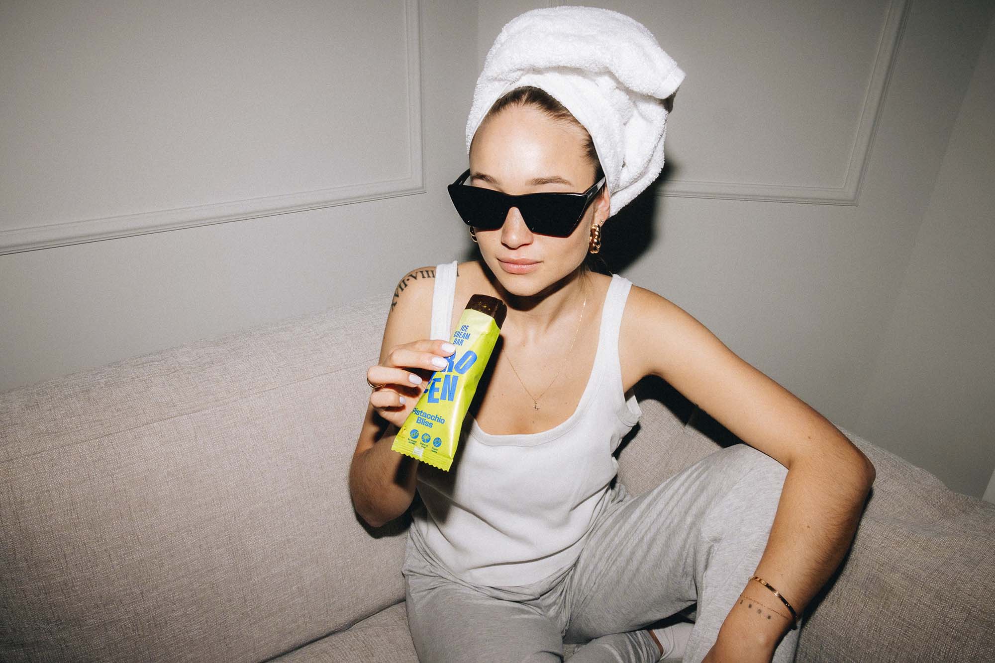 Frau mit Sonnenbrille und Handtuchturban sitzt entspannt auf einem Sofa und genießt einen Frozen Power Pistachio Bliss Eis-Riegel, vegan und ohne Zuckerzusatz.
